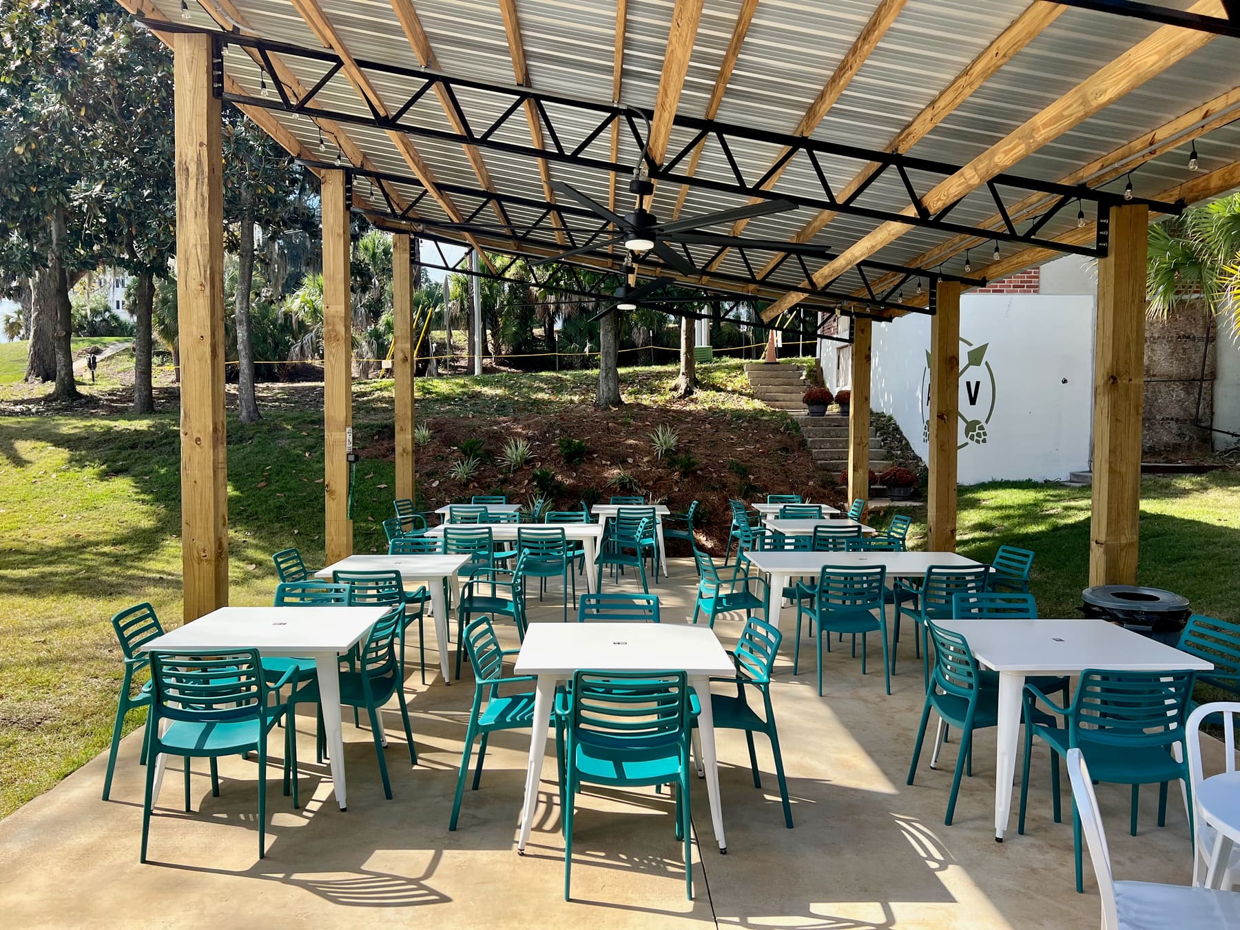 The beer garden: teal chairs and white tables on a concrete pad under a timber and steel pavilion, with lawn and palms beyond.