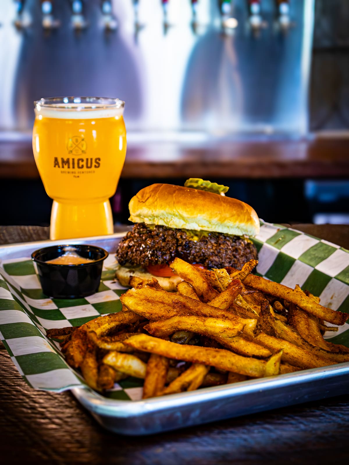 A smash burger and a pile of hand-cut fries on a green checked tray, next to a hazy beer in an Amicus glass.
