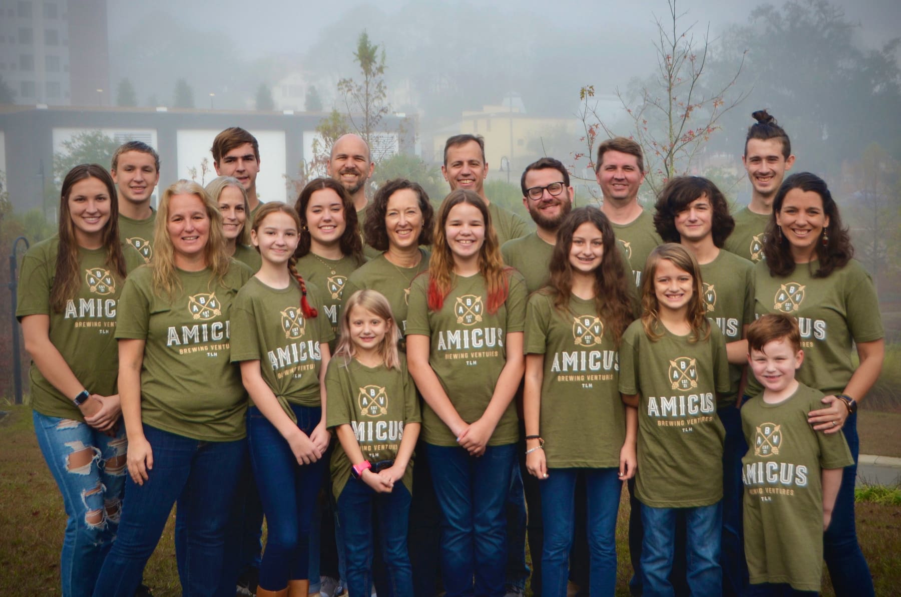 Twenty people of all ages in matching olive Amicus shirts, standing together on a misty lawn.