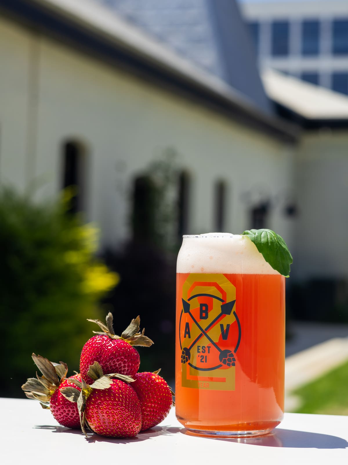 A tall glass of pink-orange beer with a basil leaf on the rim, beside four strawberries in the sun.