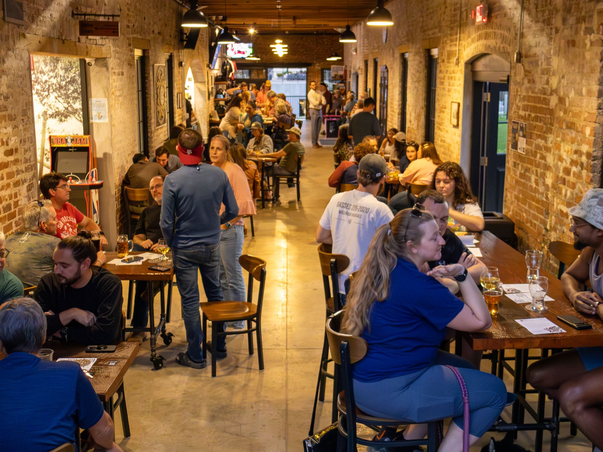 The taproom full of people at wooden tables under pendant lamps, between two long walls of bare brick.
