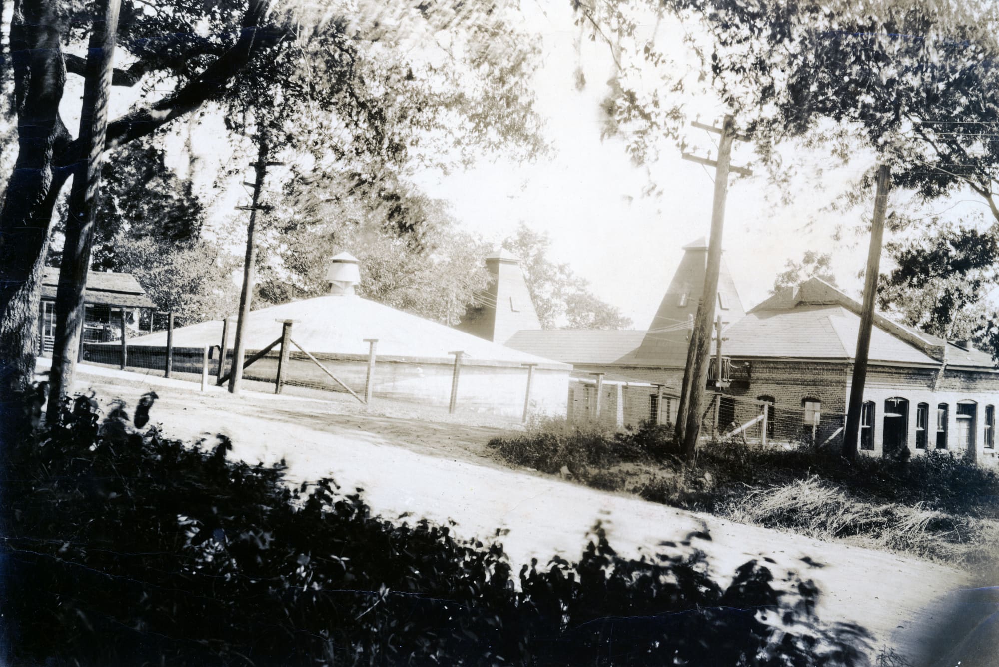 A sepia photograph of the Waterworks around 1920: a low brick building and a conical roof behind a round cistern wall, seen from a dirt road.